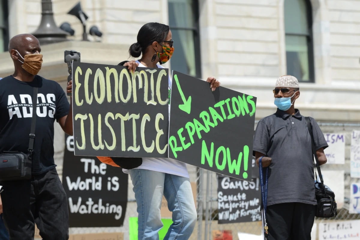 Photo of protestor holding sign that reads "Economic Justice" with an arrow pointing to another sign that reads "Reparations Now"