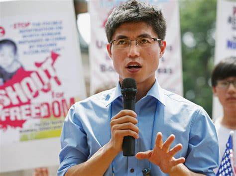 A man with glasses and a blue shirt speaks into a microphone. Behind him are signs, one reading, “Shout for Freedom.”