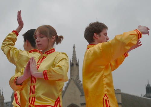 Three people in bright yellow outfits with red accents stand back-to-back while holding three different poses commonly seen in the practice of Falun Gong.