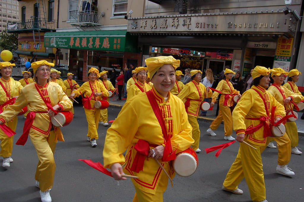 A parade of people wearing bright yellow outfits with red accents walk down the streets with drums.