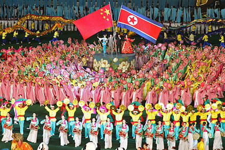A colorful festival takes place with dancers and streamers. On a pedestal, four people stand holding the flags of North Korea and China.