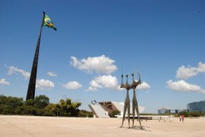 A large plaza with a statue of two elongated figures, the Brazilian flag flying, and buildings in the distance. 