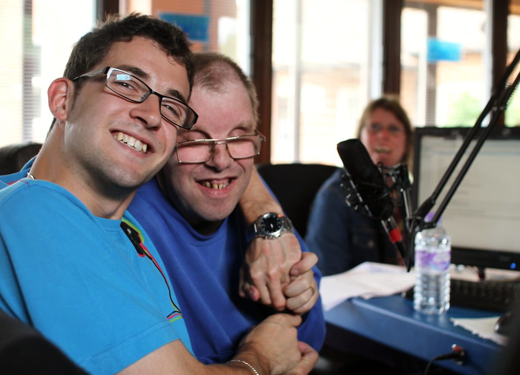 An image of two individuals with disabilities smiling at the camera in front of a microphone. 