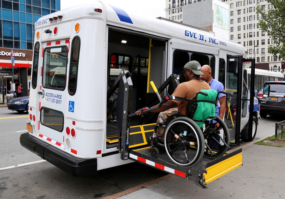 An image of an individual in a wheelchair being hoisted onto the bus, with the assistance of the driver. 