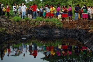 Indigenous people standing behind and looking at an open and unlined oil pit. 