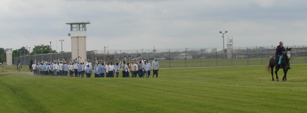 Alt text: An image depicting a line of inmates, all who look like they are people of color, each holding a shovel in hand walking in a line inside the penitentiary, as a white man rides on a horse away from them. 