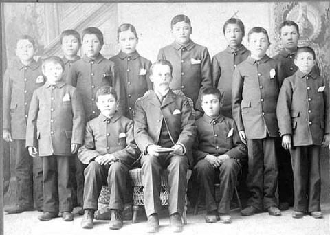 An image of indigenous children dressed in military garb posing with an adult at one of the boarding schools set up across the country in efforts to assimilate the children. 