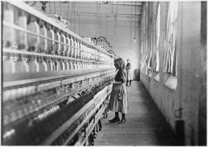 A black and white photo that shows a large textile machine with a child standing in the foreground and an older person standing blurred in the background