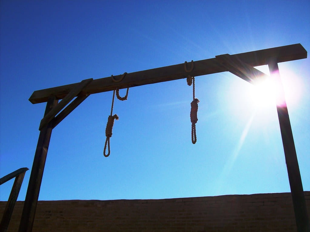 The sun shines behind a wooden gallows with two nooses hanging down. 
