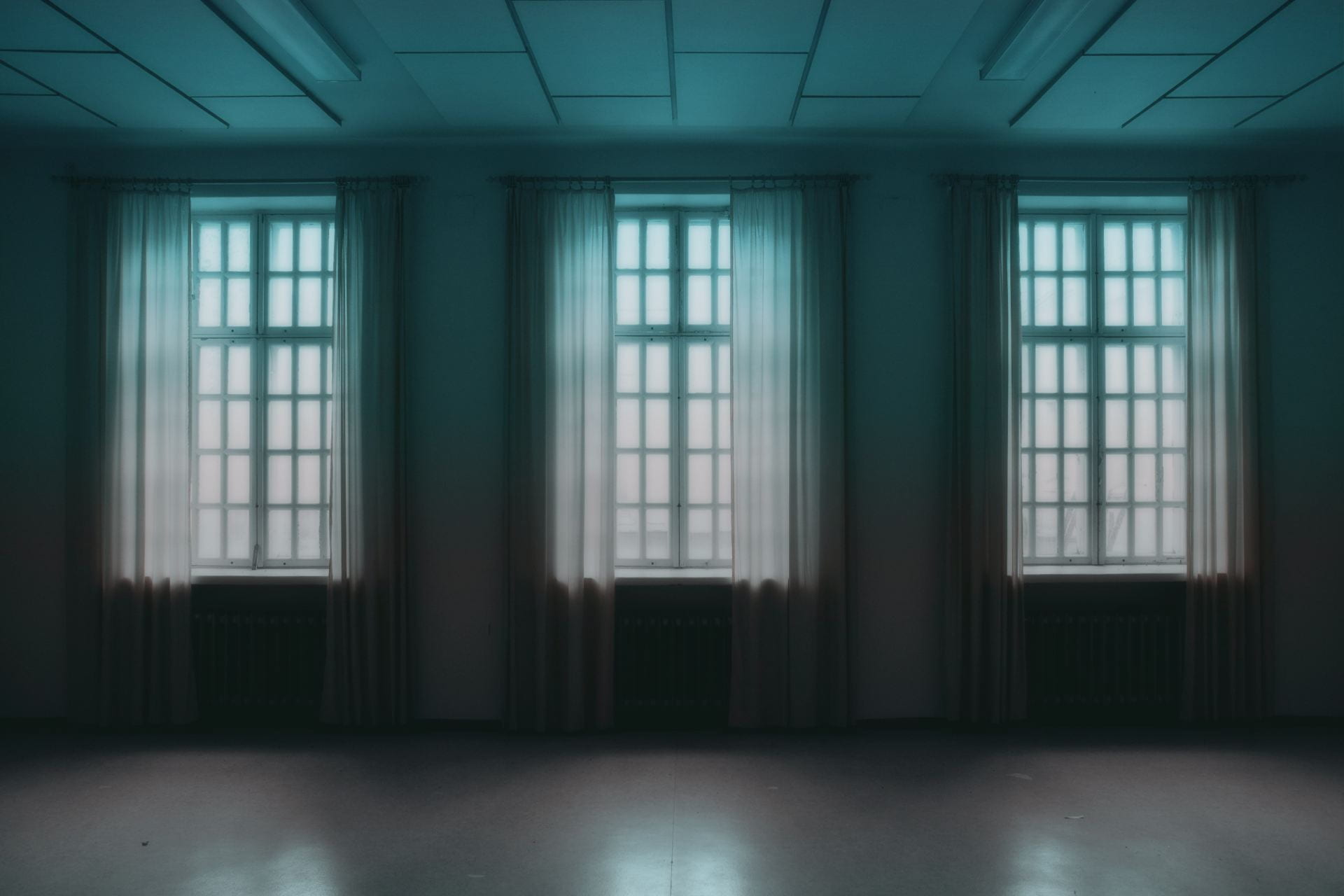 An empty room with three windows, all with long, sheer curtains. The two ceiling lights are off. Nothing but light can be seen outside the windows.