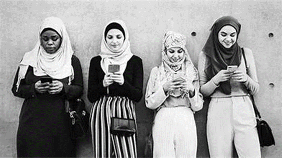 Four teen girls in hijabs paired with modern clothing are leaning against a wall, looking at their phones. Source: Yahoo Images