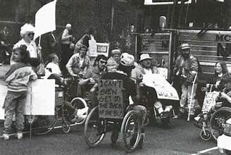 a black and white photo featuring disability rights advocates. In the center, a person in a wheelchair has a sign that reads "I can't even get to the back of the bus."