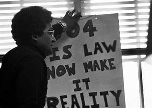 Black and white image of a person holding a protest sign that reads "504 is law now make it reality."