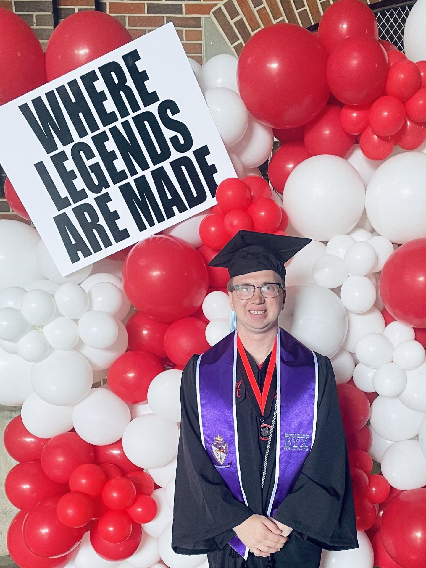 Colby stands in a black graduation cap and gown. He stands in front of a wall of red and white balloons, with a sign above that reads "where legends are made."