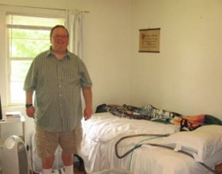 A middle-aged man stands inside a room at a group home in front of a bed. Source: Yahoo Images
