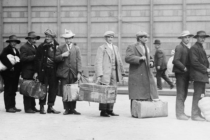 A black and white photo of men wearing clothing from the early 1900s. The men are carrying suit cases and standing in a line at Ellis Island after arriving in the US.