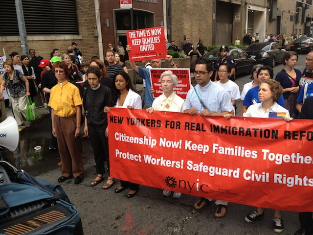 A group of protesters standing with a large red sign. The sign reads "New Yorkers for Real Immigration Reform." Underneath, it says "Citizenship Now! Keep Families Together! Protect Workers! Safeguard Civil Rights!"