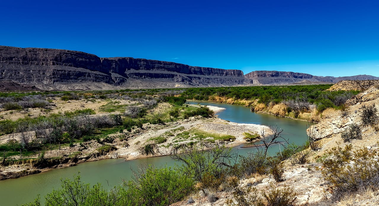 A view of a bluish green river stretching through the desert. Mountains are present in the background. The shore of the river is mostly sand, with some short green shrubbery present.