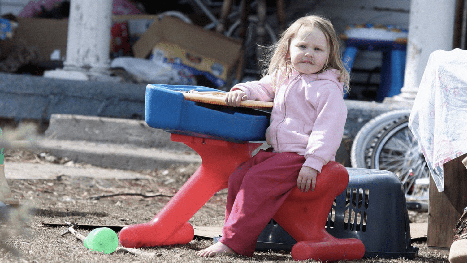 A child with no shoes, pink pants, and a light pink jacket sits on a red, blue, and yellow hard plastic toy. She is in a backyard surrounded by gray dirt, trash, and other junk. Her face is dirty, and her hair is messy.