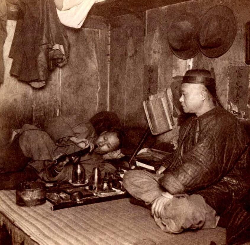 Opium users sit and lay relaxing on the floor of a small and organized Opium Den, wearing traditional Chinese clothing and smoking the drug through a pipe next to a tray of materials.