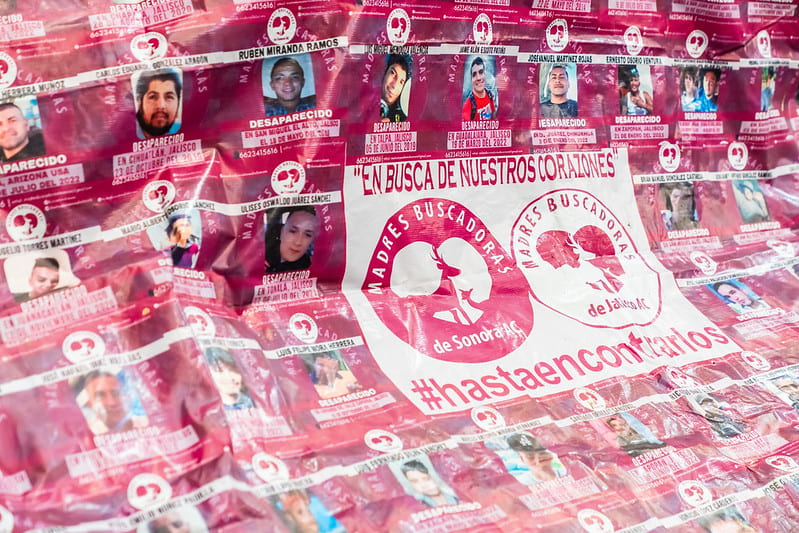 Image Description: A large pink banner with "En Busca De Nuestros Corazones" and "Madres Buscadores" written across the middle. There are many images of disappeared people. Source: https://www.flickr.com/photos/filguadalajara/53368996982/in/photolist-2pj34HD-2pj8EbH-2pja1kA-2pj34so-2pj34n8-2pj9iQb-2pja1gc-2pj34eC-2pja2pQ-2pj9ZZR-2pj9ZCt-2pj7GEv-2pj8Ept-2pj8EkA-2pj34Fu-2pj33ZQ-2pj8DJf-2pj9hM9-2pj34qz-2pja1rx-2hS7sD8-2nmdgdT-2pj8Dww-2nmdg8H-2nmdgpE-ULdzFe-UUkAHs-UXQmFc-TFsA3Q-TFswrS-UUkBAu-UXQnGF-UXQozT-TFsyG3-2nmdgiH-2nmdco6-ULdAX2-2nm7Utq-2nm7U3L-2nmdcJ6-2nmfHX7-2nmfHM2-2nm7UmM-ULdAcV-2nmeqmr